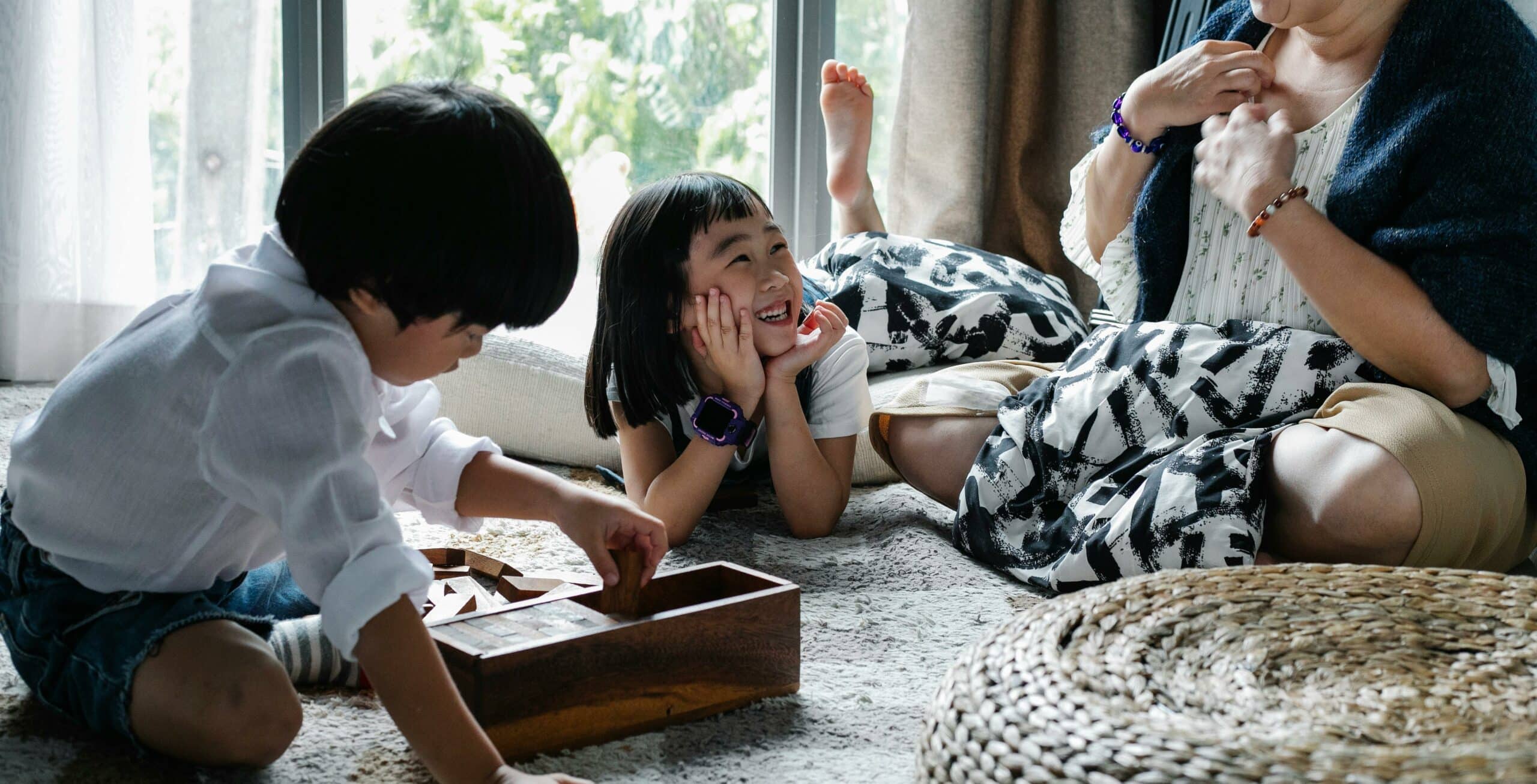 Attentive nanny and two joyful children laying on a carpet engaged in a board game, showcasing the nurturing and interactive childcare provided by Lifetime of Love nannies