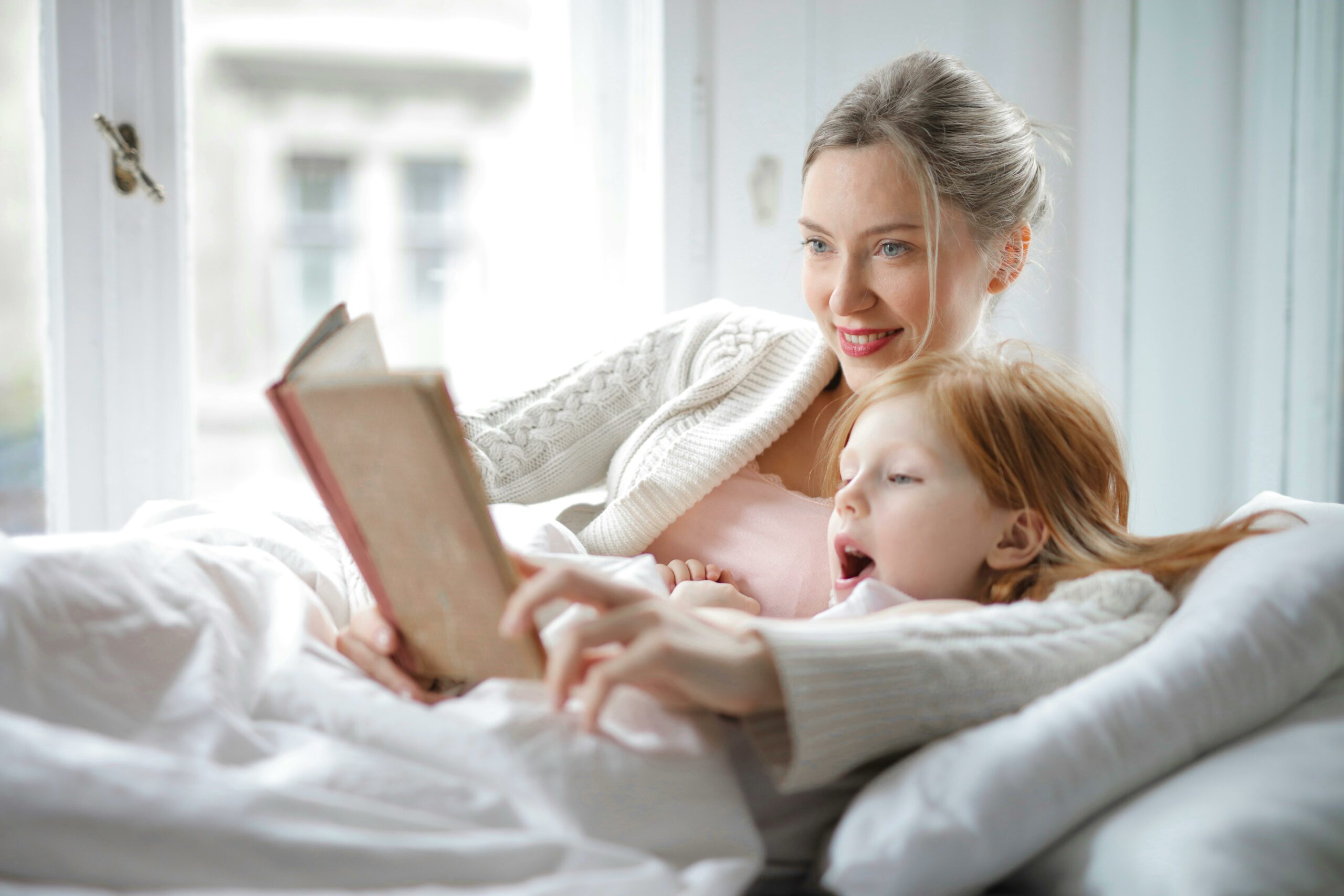 Attentive nanny and young girl enjoying a book together in bed, showcasing the experienced and reliable one off childcare services offered by Lifetime of Love Nannies