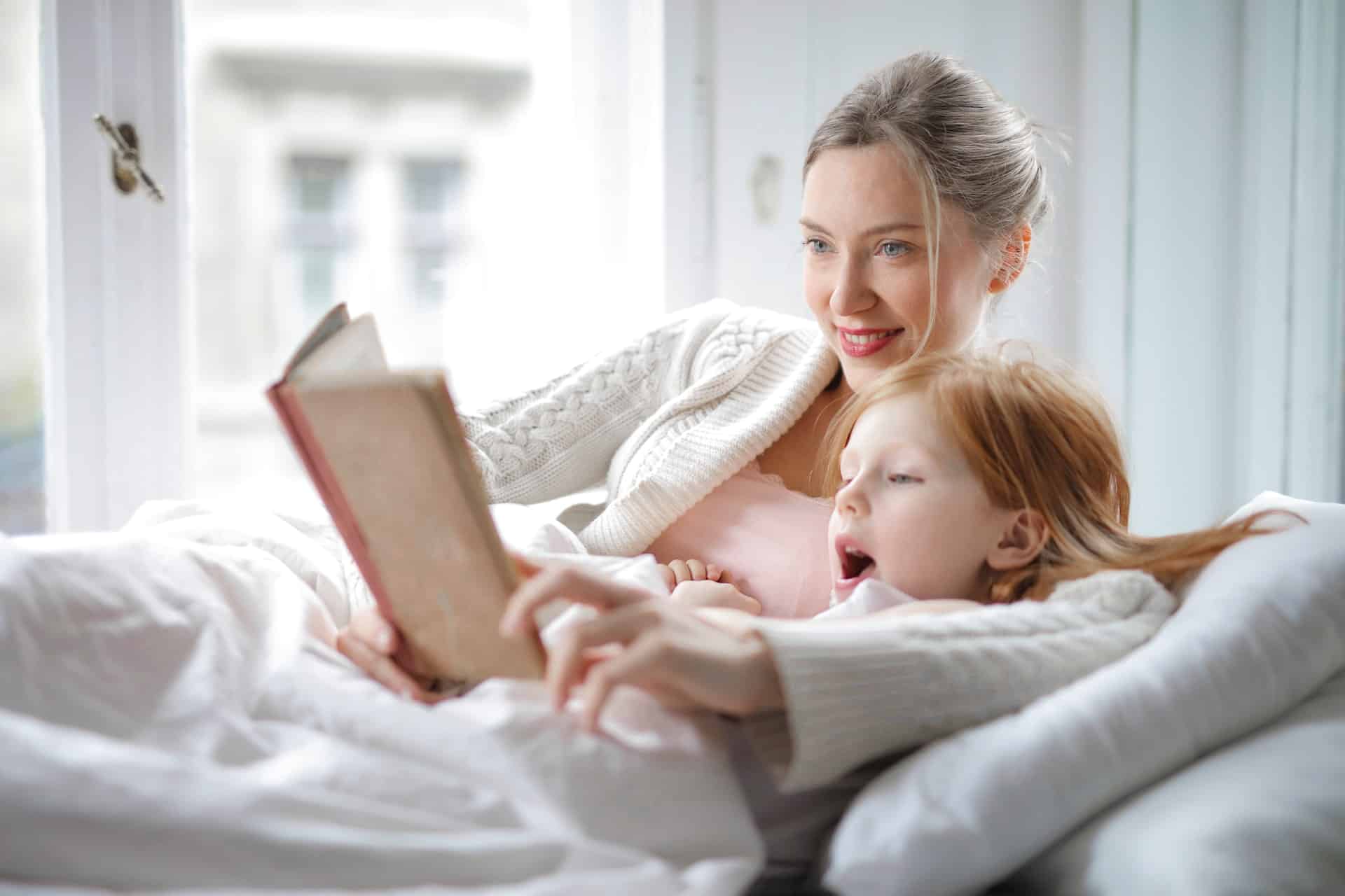 Blonde blue eyed nanny reading a bedtime story to a redhead girl both comfortably settled in bed ready for sleep