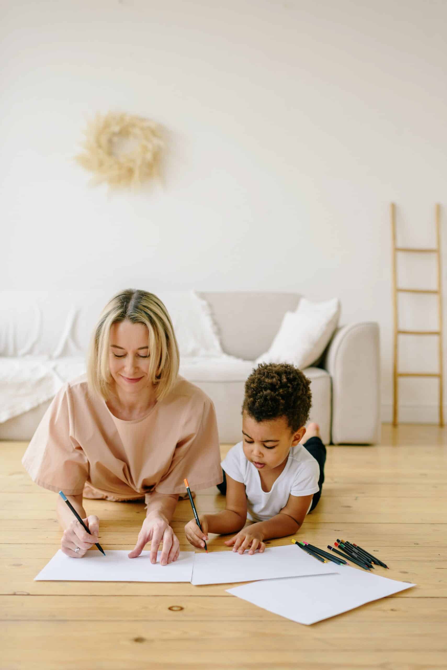 Engaged nanny and child joyfully drawing together on a wooden floor, highlighting the flexible and reliable childcare solutions provided by Lifetime of Love Nannies