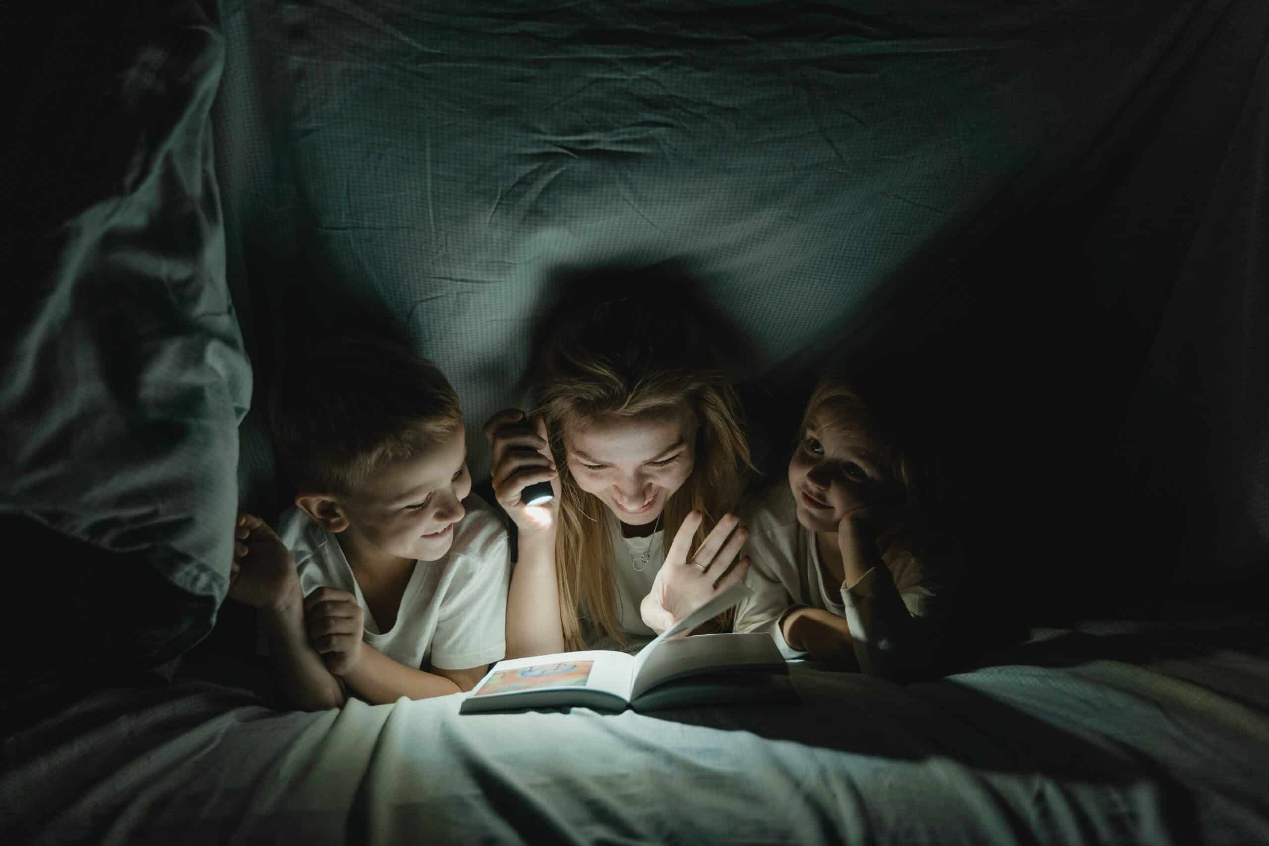 Enthusiastic nanny creating a magical reading moment under the bed sheets with two children using a flashlight, reflecting the nurturing and engaging childcare provided