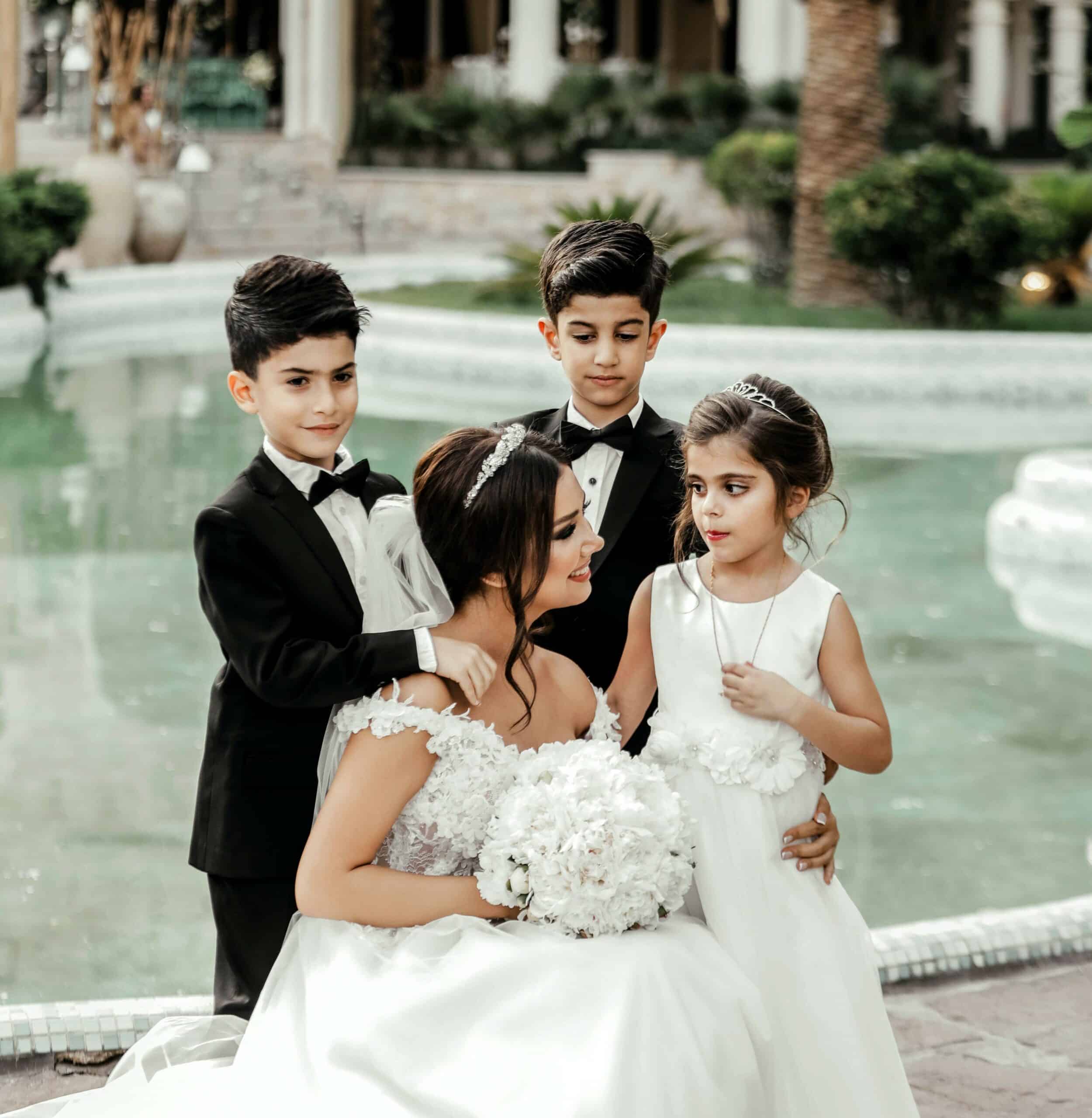 Smiling bride posing with three happy children in front of an elegant fountain at a classical wedding venue showcasing Lifetime of Love Nannies event childcare scaled
