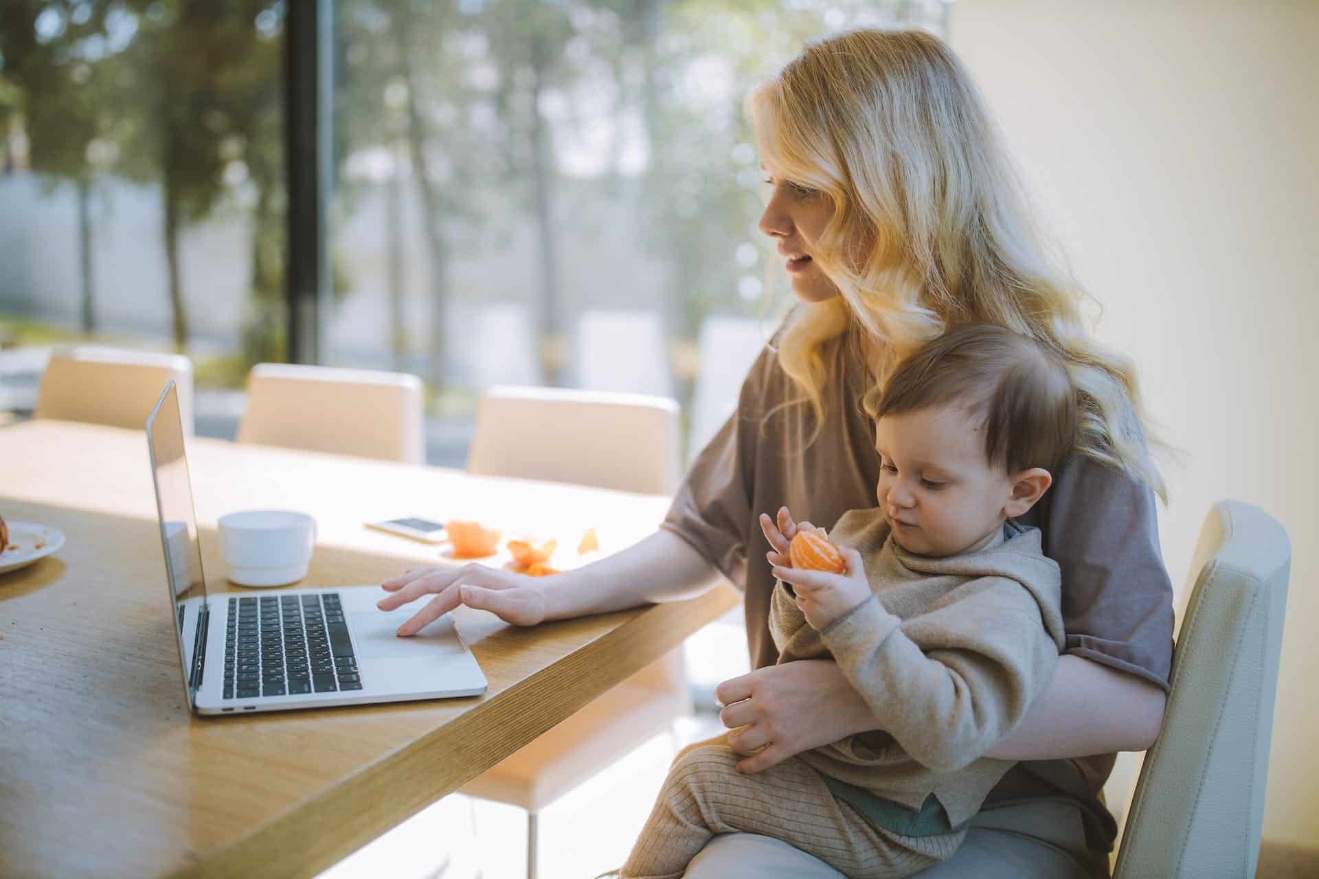 Blonde mother multitasking with work on her laptop while holding her young son, who is enjoying a tangerine
