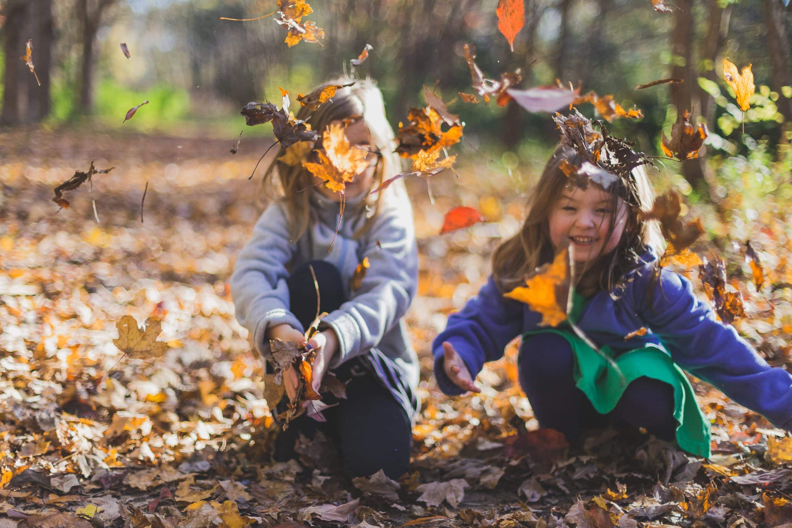 Two girls joyfully playing with autumn leaves near the woods
