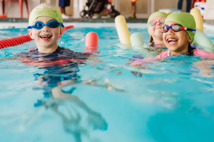 Three young children wearing swim caps and goggles smile while taking swim lessons in a pool with colorful swim noodles, supervised as part of water safety education at British Swim School