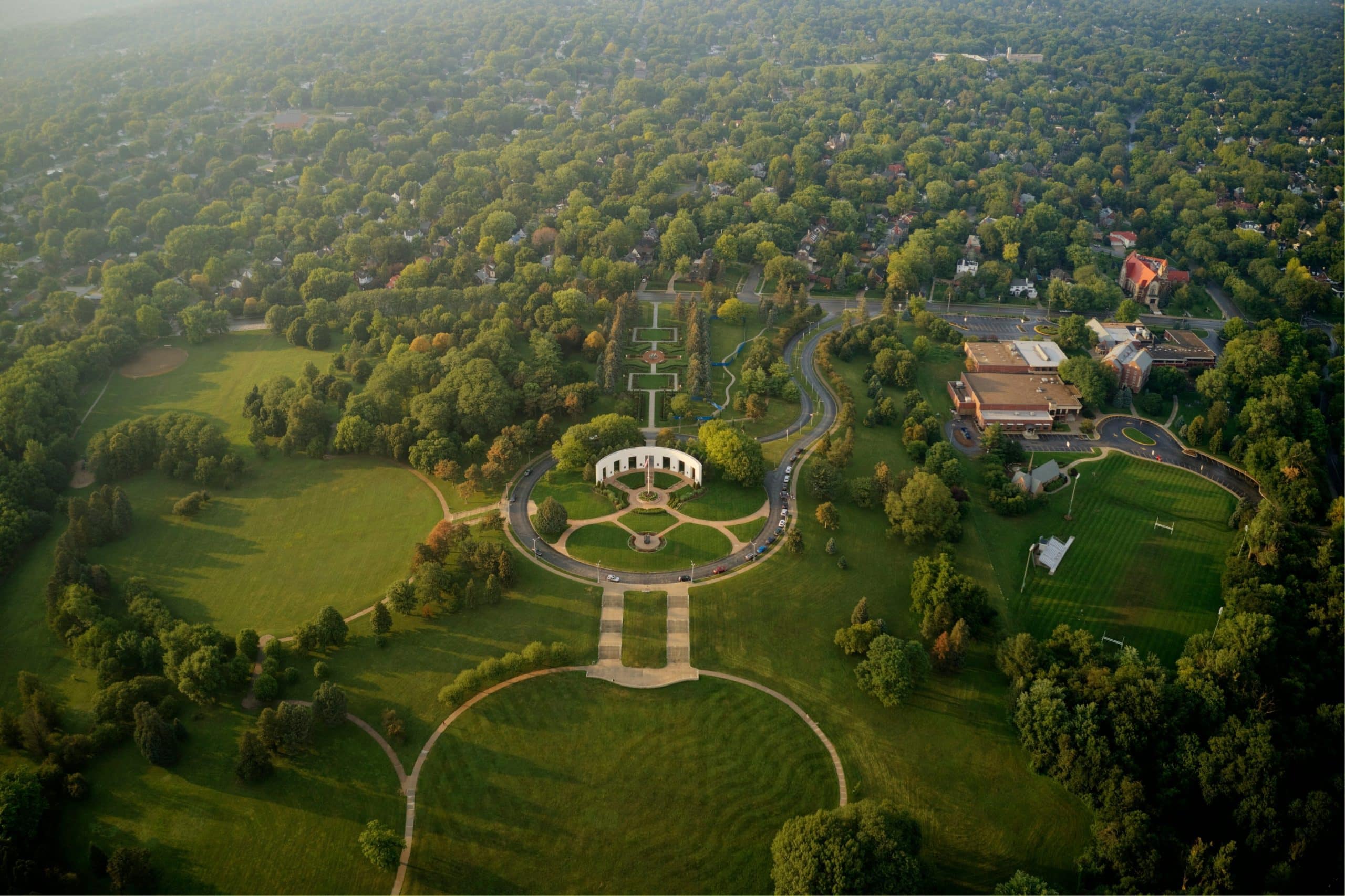 Aerial view of a tree lined Omaha park with a circular memorial plaza and walking paths near a residential neighborhood